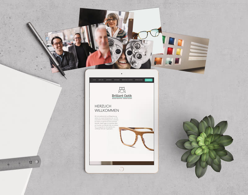 Betontisch von oben: ein iPad zeigt die Startseite von Brilliant Optik mit dem Eulen-Logo, daneben Fotos aus dem Laden.