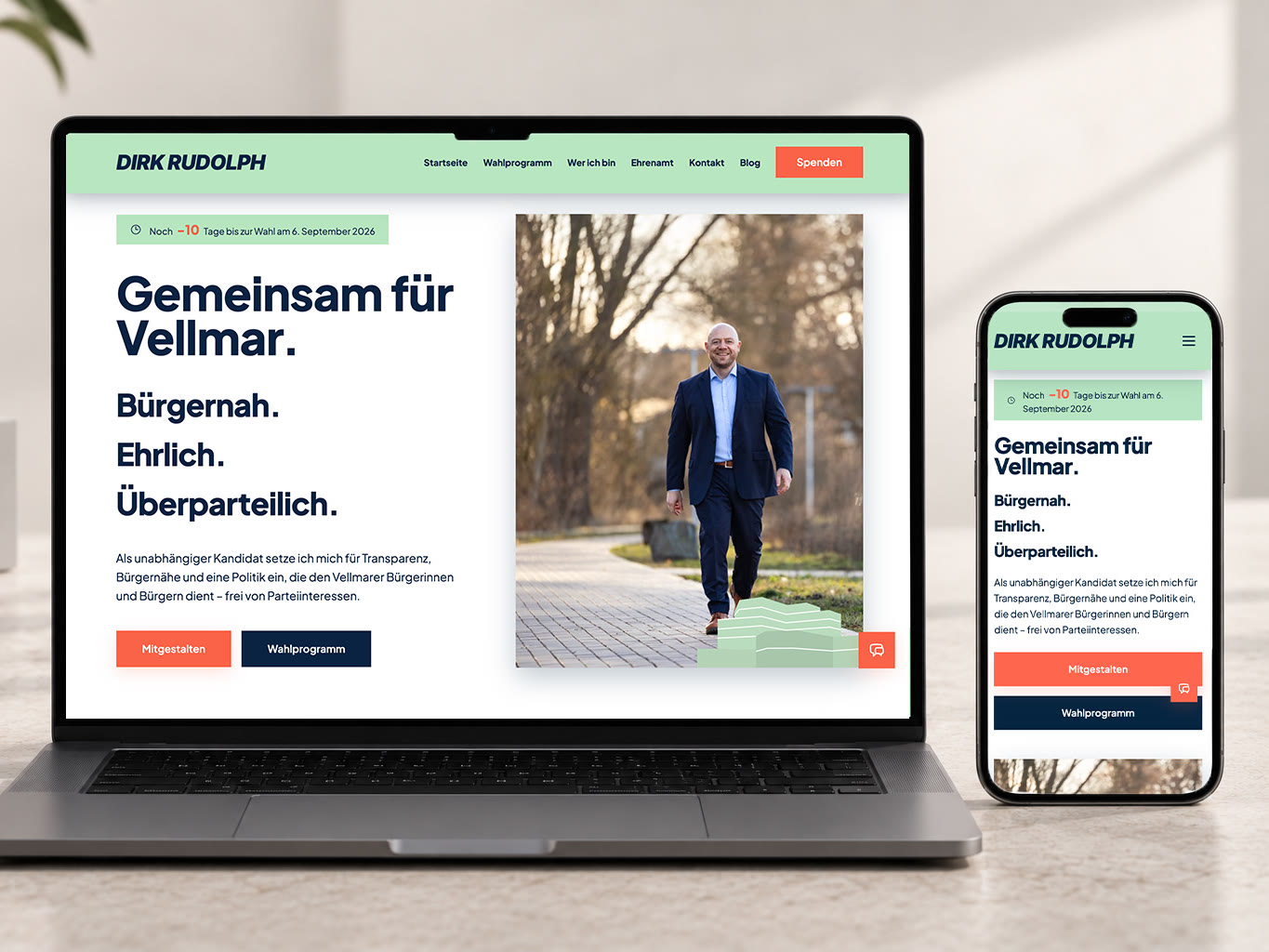 Laptop und Smartphone auf einem hellen Tisch, beide mit der Startseite von dirk-rudolph-vellmar.de: hellgrüne Navigation, die Überschrift „Gemeinsam für Vellmar.“ und die Zeilen Bürgernah. Ehrlich. Überparteilich., daneben ein Foto von Dirk Rudolph, der auf die Kamera zugeht.