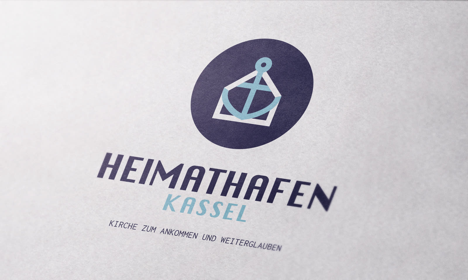 Das Heimathafen-Logo auf mattem Papier: in einer dunkelblauen Ellipse ein hellblauer Anker, der zugleich ein Haus umschreibt und in dem ein Kreuz steckt. Darunter „HEIMATHAFEN Kassel“.