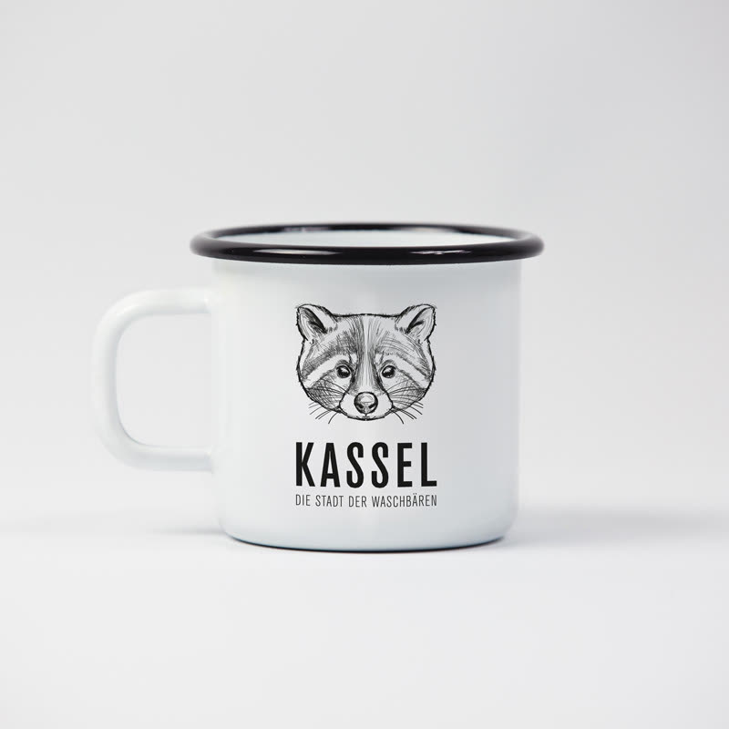 Weiße Emailletasse mit schwarzem Rand: darauf ein fein gezeichneter Waschbärkopf, darunter „Kassel – die Stadt der Waschbären“.