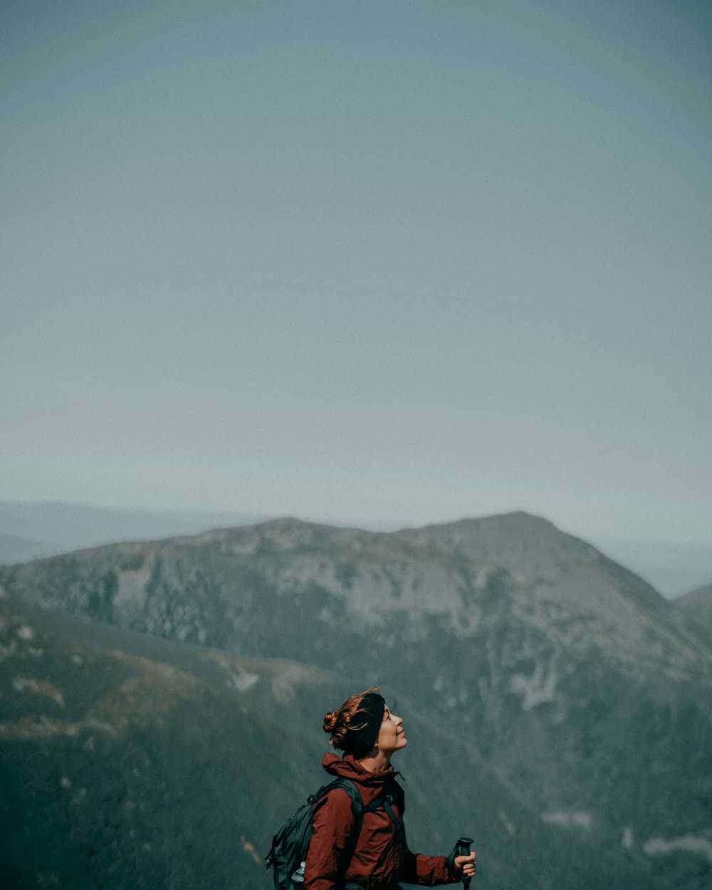 Eine Frau in roter Jacke steht mit Rucksack und Wanderstock auf einem Bergrücken und schaut nach oben. Hinter ihr liegen weitere Höhenzüge im Dunst, darüber ein weiter grau-blauer Himmel.