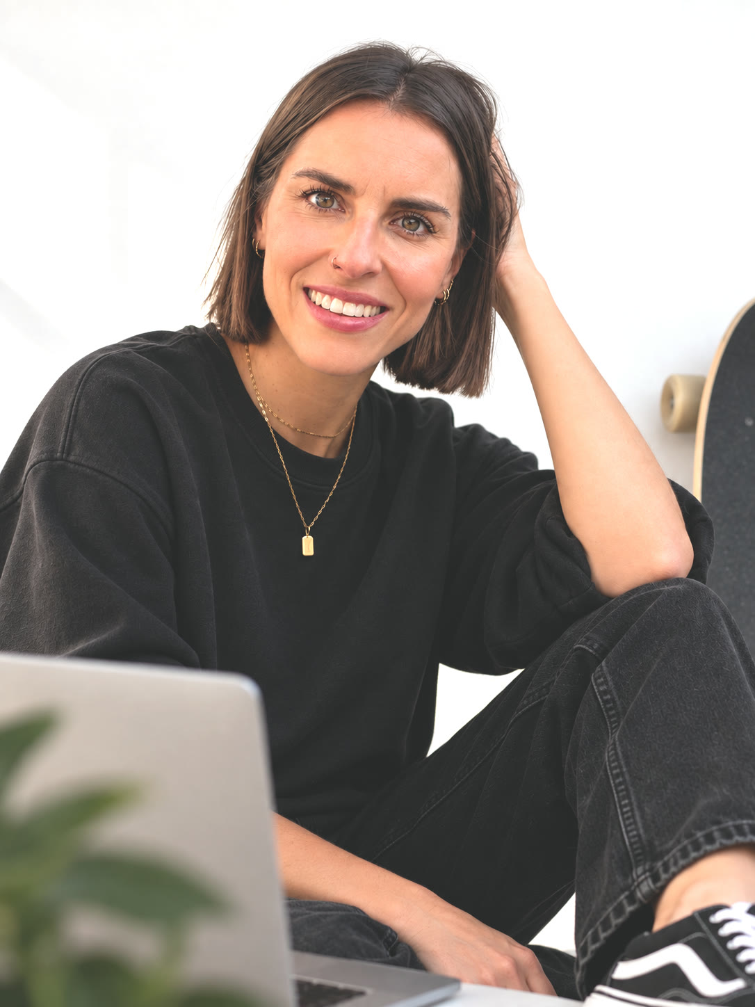 Julia Reuter sitzt vor einer hellen Wand auf dem Boden, den Blick in die Kamera: dunkler Bob, schwarzes Sweatshirt, schwarze Jeans, eine Hand im Haar, ein offenes Lächeln. Neben ihr ein aufgeklappter Laptop, ein Skateboard und eine Grünpflanze.